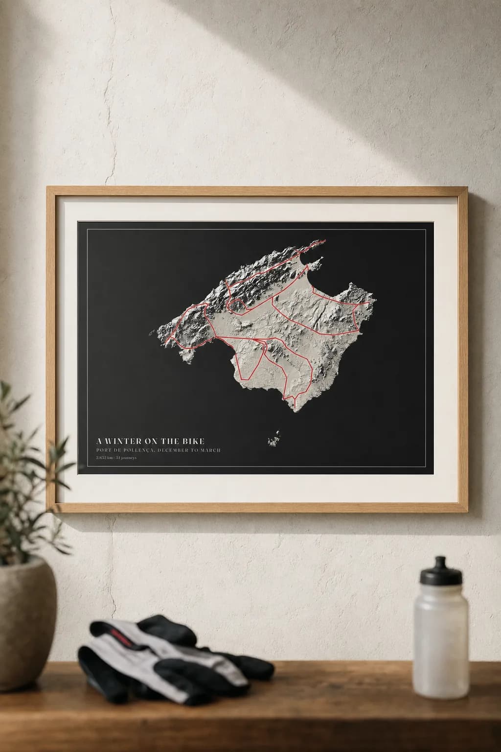 A poster of a winter of riding on Mallorca in plaster relief — the island carved in pale light and shadow on black paper — framed in oak above a console with cycling gloves