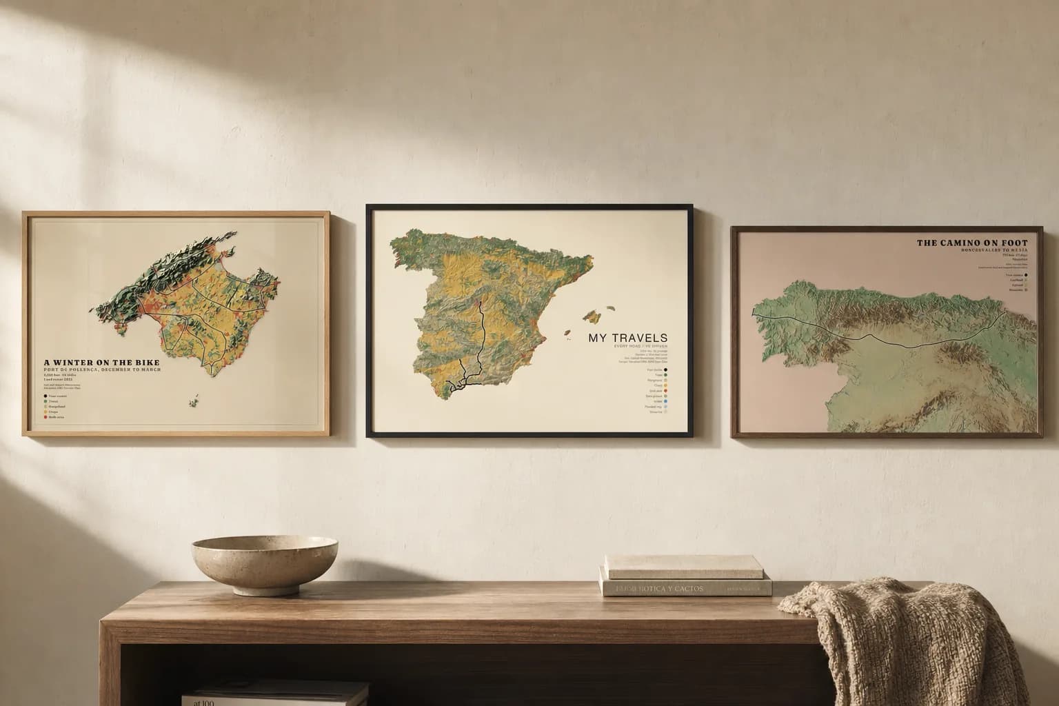 Three posters hung as one row on a hallway wall: a Mallorca cycling map on sand paper in oak, a Spanish road trip on cream in black, and the Camino on clay-pink in walnut.