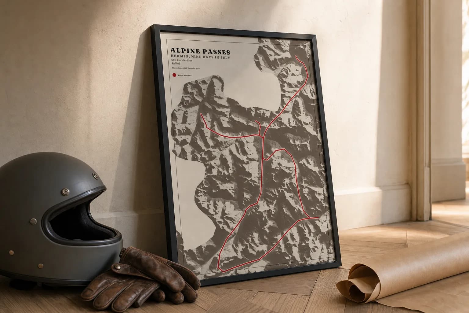 The framed Alpine passes poster on bone paper leaning against a hallway wall on a herringbone floor, a motorcycle helmet, leather gloves and the kraft wrapper beside it.