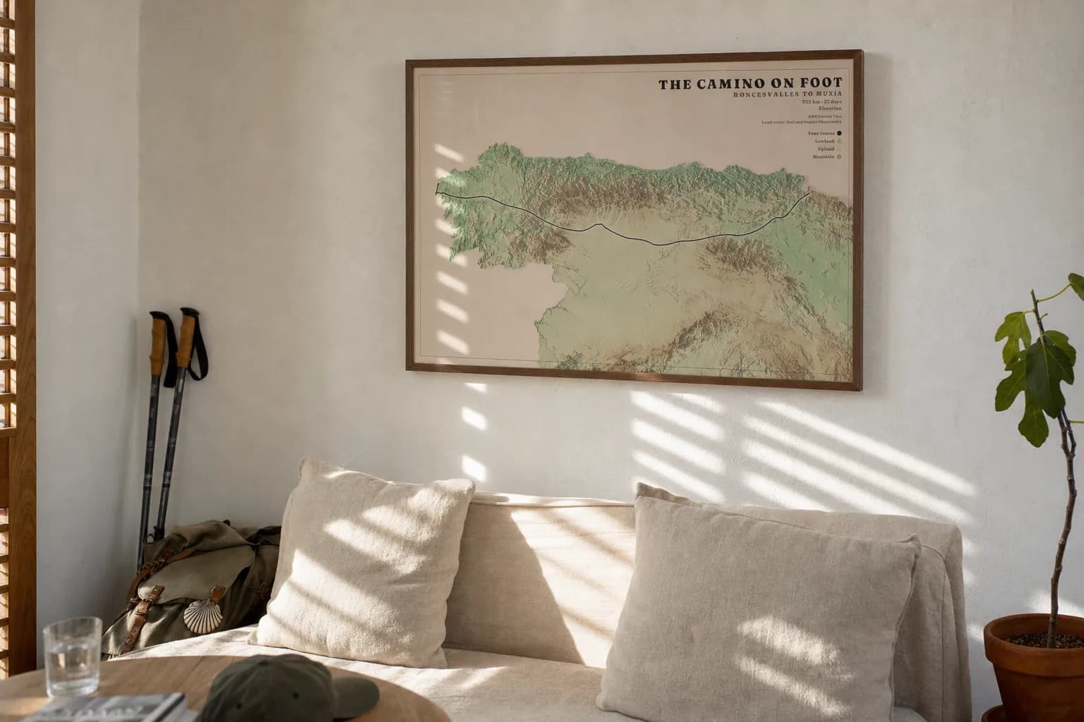 The Camino across northern Spain on clay-pink paper, in a walnut frame above a low linen sofa in a small studio flat, midday sun through timber shutters striping the wall.