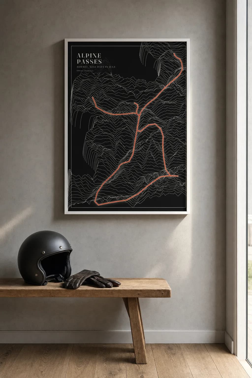 Nine days riding the passes around Bormio drawn as Ridgelines — cream lines rising into the Alps on black paper, an orange route over them — in a white-painted frame above a wooden bench with a motorcycle helmet and gloves.